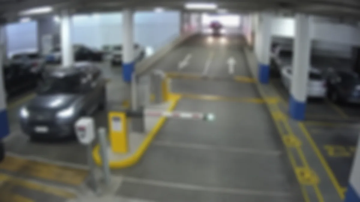 Blur degradation in a car park CCTV view, with vehicles and barrier details smeared and unreliable.