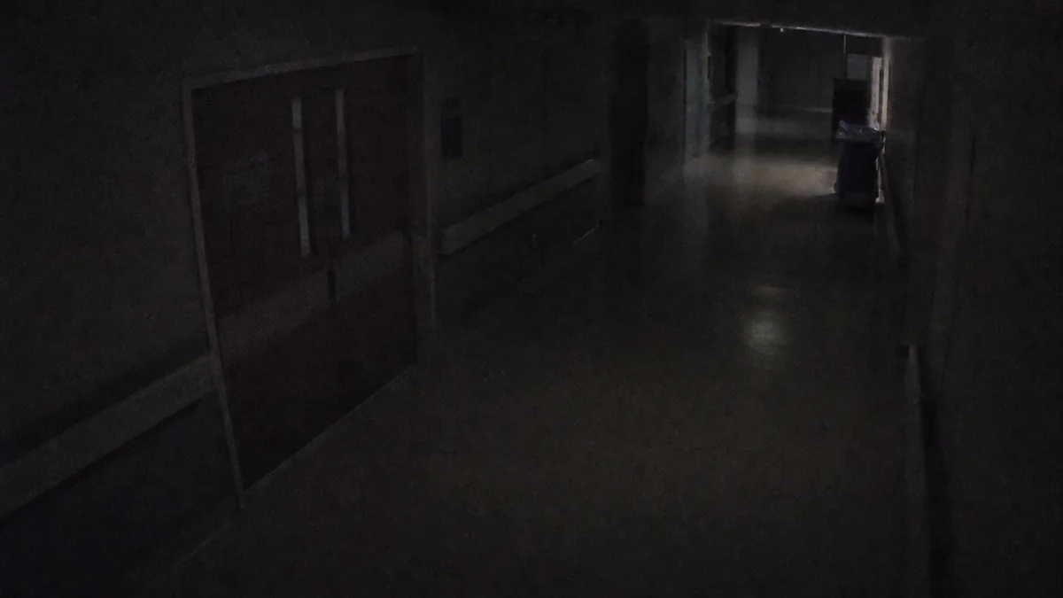 Darkness degradation in an institutional corridor CCTV view with weak emergency lighting and noisy shadows.