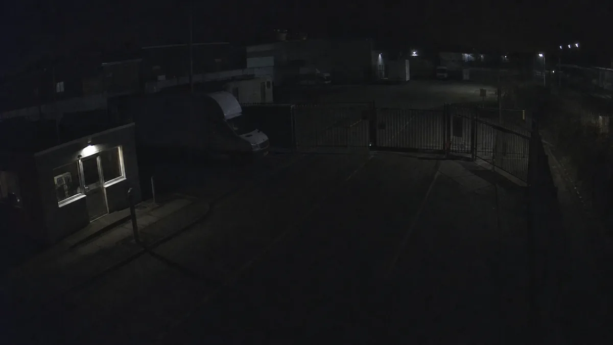 Darkness degradation in a logistics yard CCTV view with only silhouettes visible at night.
