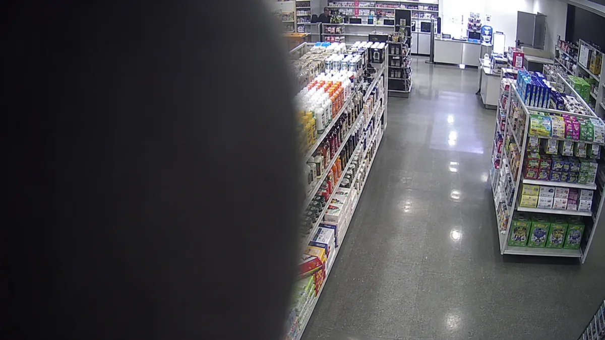 Occlusion degradation in a retail aisle CCTV view, with a dark object blocking the left side of the lens.