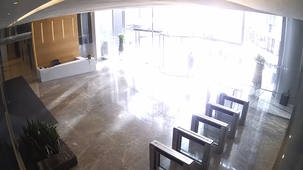 Overexposure degradation in a corporate atrium CCTV view, with the glass entrance and floor reflections clipped white.