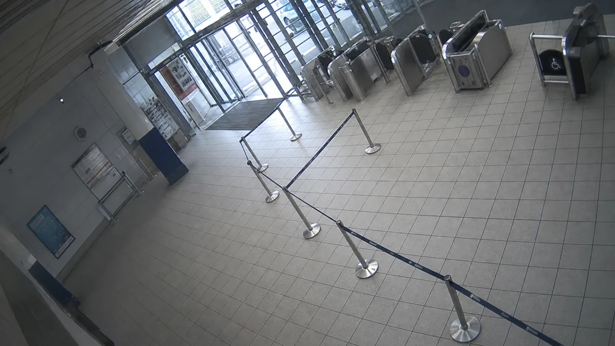 Tilt degradation in a transport concourse CCTV view, with tile grid and queue barriers off level.